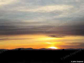 Ridge Sunset – Sun sinking behind a distant ridge under layered clouds.