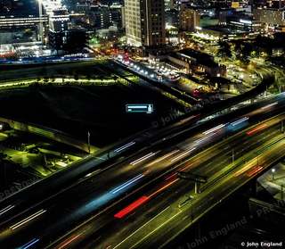 Freeway Light Streams at Night – Aerial long-exposure-style freeway light trails beside downtown towers.