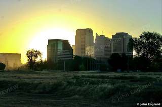 Sunrise Behind the Towers – The sun blazing behind downtown towers over an open field. John England.
